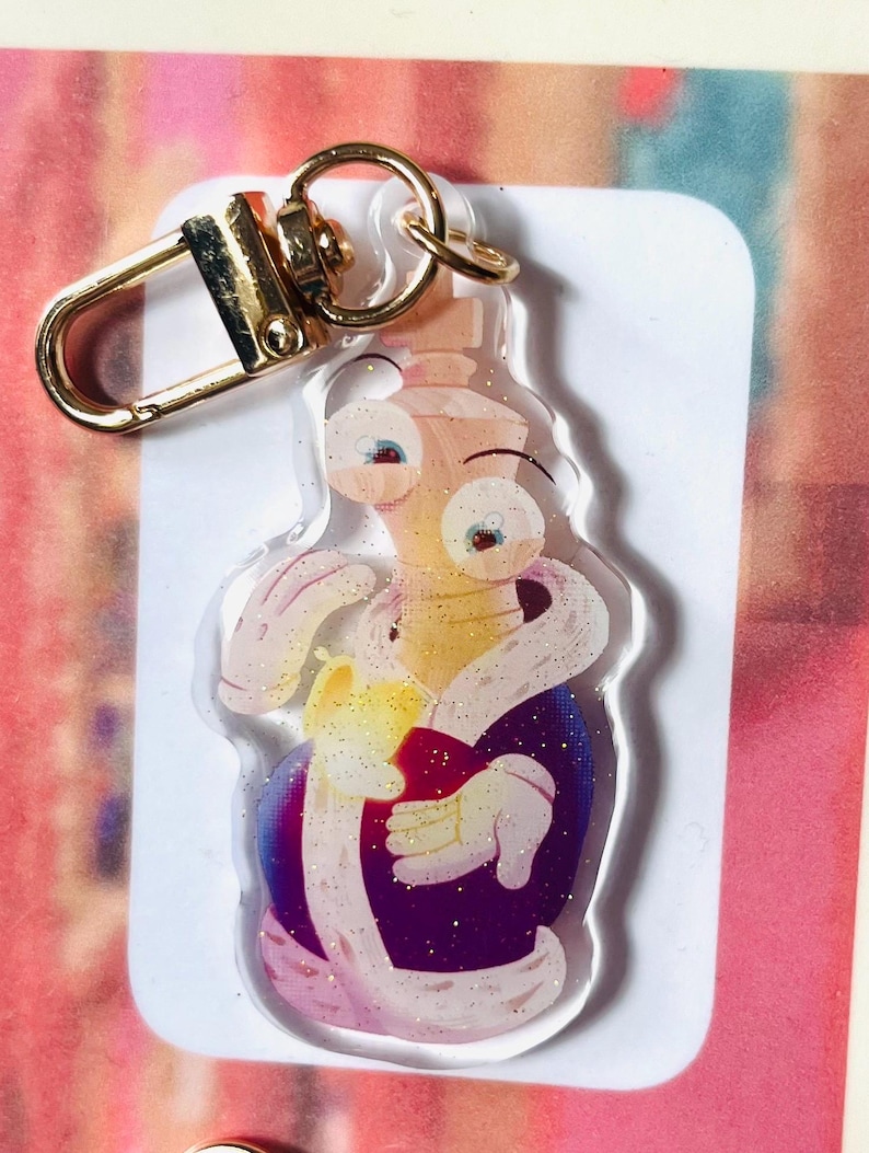 May include: A clear acrylic keyring featuring a cartoon character in a purple robe holding a red object. The keyring has a gold-coloured clasp and ring. The character has large eyes and is surrounded by glitter.