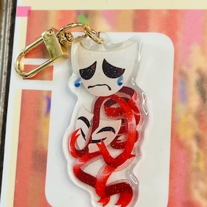 May include: A clear acrylic keyring featuring a sad theatre mask with blue tears and a red ribbon design. The keyring has a gold-coloured clasp and a white background. The word "CHOOSE" is visible in the background.