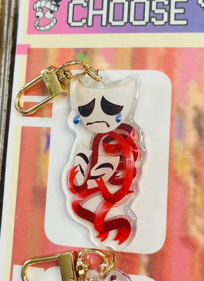 May include: A clear acrylic keyring featuring a sad theatre mask with blue tears and a red ribbon design. The keyring has a gold-coloured clasp and a white background. The word "CHOOSE" is visible in the background.