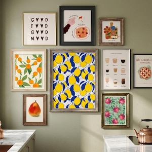A gallery wall displaying nine framed kitchen-themed art prints including lemons, coffee, pomegranates, and pie against a pale green wall above a kitchen counter.
