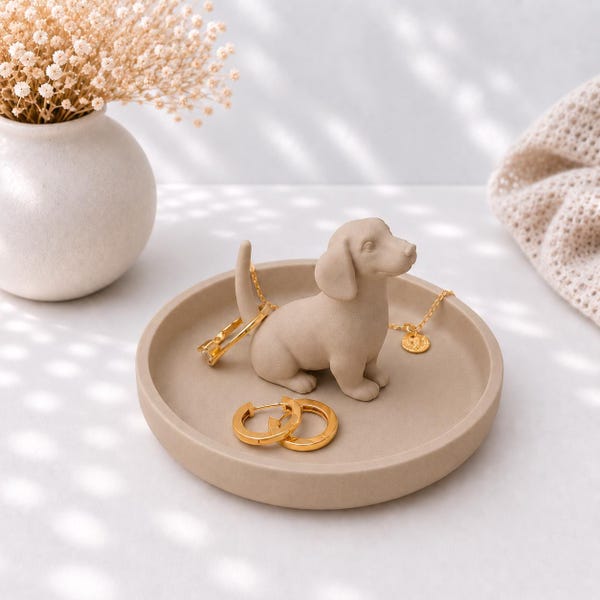Könnte beinhalten: Eine beige, kreisförmige Schmuckschale mit einer kleinen, geformten Dackel-Figur in der Mitte. Die Schale enthält eine goldene Halskette mit einem kleinen runden Anhänger sowie ein Paar passende goldene Creolen, die auf der glatten Oberfläche liegen.