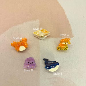 Op de afbeelding: Een verzameling van vijf miniatuur bedels met oceaanthema. Stijlen zijn onder meer een oranje krab, een zeeschelp, een zeepaardje, een paarse octopus en een blauwe walvis. Elke bedel heeft een uniek ontwerp en is felgekleurd. De tekst "Style A" tot "Style E" is inbegrepen.