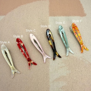 Six glossy, fish-shaped hair clips arranged diagonally on a textured pastel background. The clips feature a minimalist hollow cutout design. Colors from left to right include milky white pearl, red tortoiseshell, soft blush pink, classic brown tortoiseshell, mint green pearl, and yellow-orange marbled. Elegant coastal aesthetic and mermaidcore hair accessories for women.
