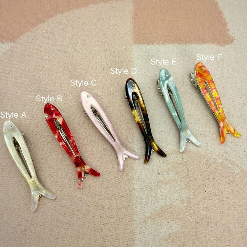 Six glossy, fish-shaped hair clips arranged diagonally on a textured pastel background. The clips feature a minimalist hollow cutout design. Colors from left to right include milky white pearl, red tortoiseshell, soft blush pink, classic brown tortoiseshell, mint green pearl, and yellow-orange marbled. Elegant coastal aesthetic and mermaidcore hair accessories for women.