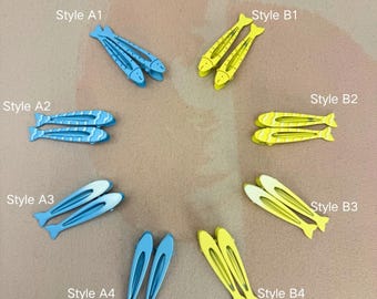 May include: A collection of eight fish-shaped hair clips arranged in pairs, labeled Style A1 through A4 in blue and Style B1 through B4 in yellow. The clips feature various patterns including solid colors, stripes, and gradients, all shaped like small fish.