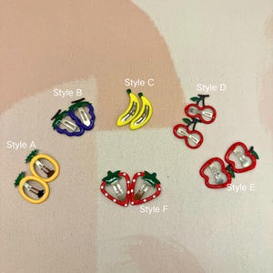Könnte beinhalten: Eine Sammlung bunter Haarspangen in verschiedenen Fruchtformen, darunter Bananen, Erdbeeren, Kirschen und Äpfel. Jede Spange hat einen Metallverschluss und ist in verschiedenen Farben umrandet. Das Bild enthält Textbezeichnungen für jeden Stil.