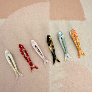 Six glossy, fish-shaped hair clips arranged diagonally on a textured pastel background. The clips feature a minimalist hollow cutout design. Colors from left to right include milky white pearl, red tortoiseshell, soft blush pink, classic brown tortoiseshell, mint green pearl, and yellow-orange marbled. Elegant coastal aesthetic and mermaidcore hair accessories for women.