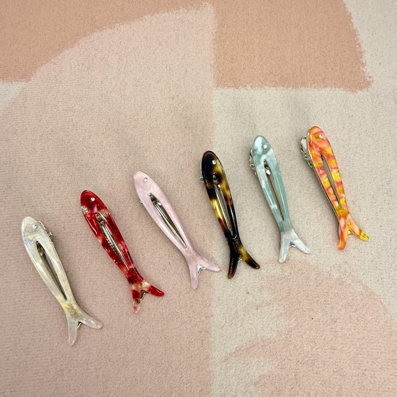 Six glossy, fish-shaped hair clips arranged diagonally on a textured pastel background. The clips feature a minimalist hollow cutout design. Colors from left to right include milky white pearl, red tortoiseshell, soft blush pink, classic brown tortoiseshell, mint green pearl, and yellow-orange marbled. Elegant coastal aesthetic and mermaidcore hair accessories for women.