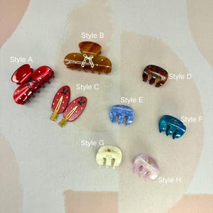 Può includere: Una collezione di otto fermagli e forcine per capelli di vari stili e colori. Gli stili includono rosso, marrone, blu e rosa. Alcuni hanno elementi decorativi come fiocchi e pois. Le clip sono disposte su una superficie rosa chiaro.