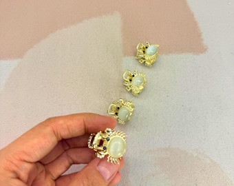 May include: Gold-toned crab rings with a central oval-shaped, off-white stone and small black and blue accent stones. The rings have intricate details and are displayed on a pink and white surface. One ring is held by a hand.