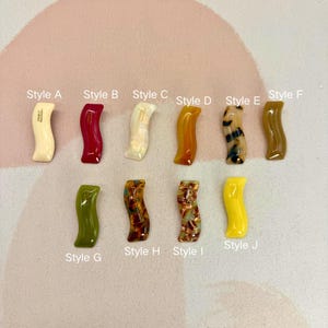 Könnte beinhalten: Eine Sammlung von zehn wellenförmigen, dekorativen Haarspangen in verschiedenen Farben und Mustern. Die Stile reichen von einfarbigen Farben wie Gelb und Grün bis hin zu marmorierten und Schildpatt-Designs. Jede Spange ist mit einem Stilbuchstaben versehen.