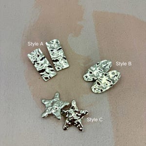 May include: Three pairs of silver-toned earrings in different styles. Style A is rectangular, Style B is oval, and Style C is starfish-shaped. Each earring has a hammered texture. The earrings are displayed on a light pink surface.