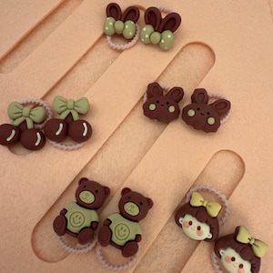 Puede incluir: Surtido de gomas para el pelo con elementos decorativos. La colección incluye gomas con orejas de conejo, cerezas, lazos, osos de peluche y caras de niña. Los colores son principalmente marrón, beige y verde claro.