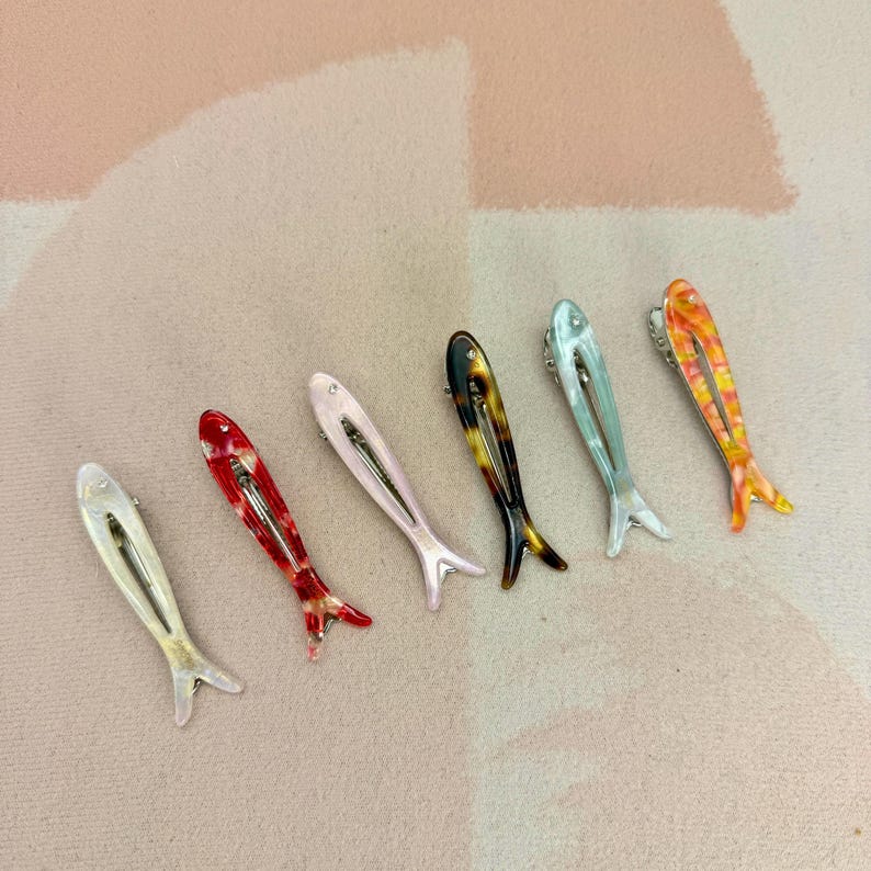 Six glossy, fish-shaped hair clips arranged diagonally on a textured pastel background. The clips feature a minimalist hollow cutout design. Colors from left to right include milky white pearl, red tortoiseshell, soft blush pink, classic brown tortoiseshell, mint green pearl, and yellow-orange marbled. Elegant coastal aesthetic and mermaidcore hair accessories for women.
