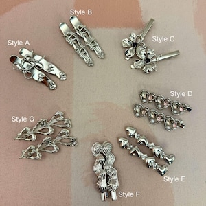 May include: A collection of silver-toned hair clips in various styles. Styles include clips with bows, floral designs, hearts, and abstract shapes. The clips are arranged on a light pink surface, showcasing their intricate details and designs.