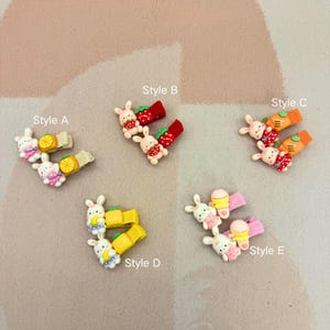 May include: Five pairs of decorative hair clips are displayed. Each clip features a small, white bunny figure with food-themed accessories, such as strawberries, carrots, lemons, and ice cream. The clips are in various colours, including red, orange, yellow, and pink.