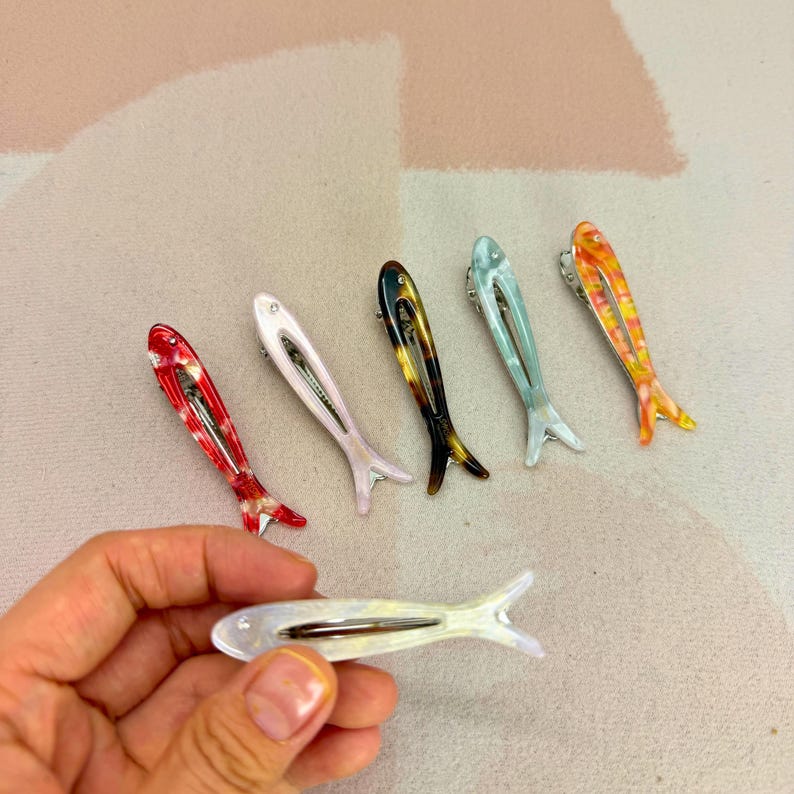 Six glossy, fish-shaped hair clips arranged diagonally on a textured pastel background. The clips feature a minimalist hollow cutout design. Colors from left to right include milky white pearl, red tortoiseshell, soft blush pink, classic brown tortoiseshell, mint green pearl, and yellow-orange marbled. Elegant coastal aesthetic and mermaidcore hair accessories for women.