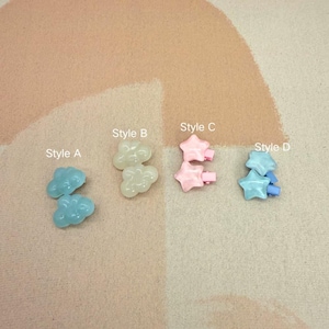 Op de afbeelding: Vier sets haarclips in verschillende stijlen. Stijl A heeft twee lichtblauwe wolkvormige clips. Stijl B heeft twee off-white wolkenclips. Stijl C bevat twee roze sterclips. Stijl D toont twee lichtblauwe sterclips.