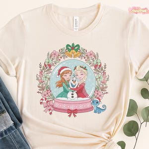 May include: Cream-colored t-shirt featuring a snow globe design with two cartoon characters, a snowman, and a small blue dragon. The snow globe is surrounded by a floral wreath with Christmas accents. The shirt has a round neckline and short sleeves.