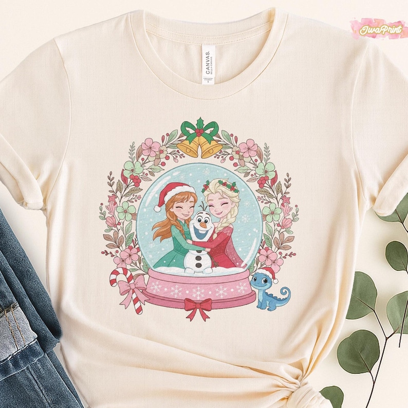 May include: Cream-colored t-shirt featuring a snow globe design with two cartoon characters, a snowman, and a small blue dragon. The snow globe is surrounded by a floral wreath with Christmas accents. The shirt has a round neckline and short sleeves.