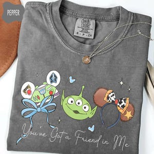 May include: A gray t-shirt featuring cartoon graphics of characters from a popular animated film. The design includes a green alien, Buzz Lightyear, and Mickey Mouse ears. The text "You've Got a Friend in Me" is written in a script font.