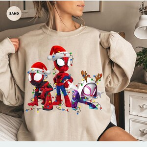 Peut inclure: Sweat-shirt beige avec trois personnages de Spider-Man en tenues de No&euml;l. Les personnages portent des bonnets de P&egrave;re No&euml;l et sont entour&eacute;s de guirlandes lumineuses color&eacute;es. Le design comprend Spider-Man, Spider-Man accroupi et Spider-Gwen.