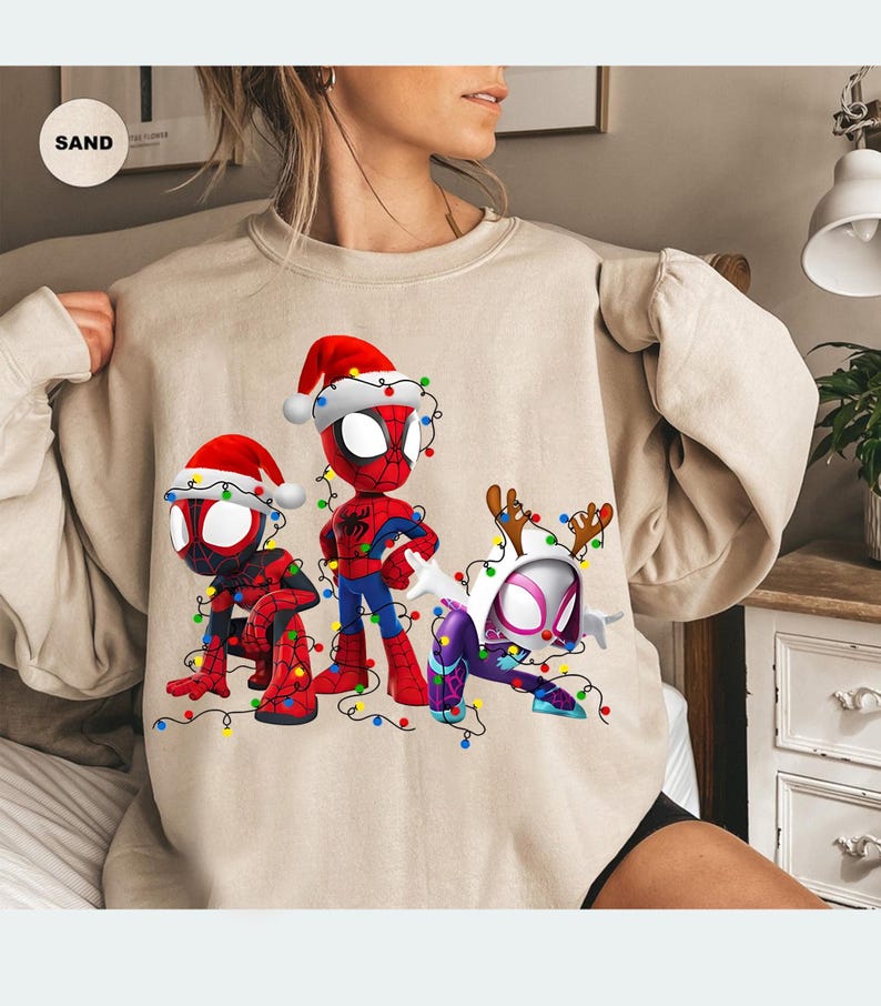 Peut inclure: Sweat-shirt beige avec trois personnages de Spider-Man en tenues de No&euml;l. Les personnages portent des bonnets de P&egrave;re No&euml;l et sont entour&eacute;s de guirlandes lumineuses color&eacute;es. Le design comprend Spider-Man, Spider-Man accroupi et Spider-Gwen.