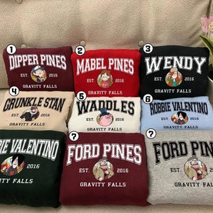 May include: A collection of sweatshirts in various colors, each featuring a different character name from the animated series "Gravity Falls." The names are displayed in a collegiate font, accompanied by character illustrations and the text "EST. 2016 GRAVITY FALLS."