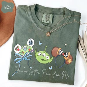 May include: Moss green t-shirt featuring a graphic of Disney characters, including Buzz Lightyear, an alien, and Mickey Mouse ears. The phrase "You've Got a Friend in Me" is written in cursive. A silver necklace is draped across the shirt.