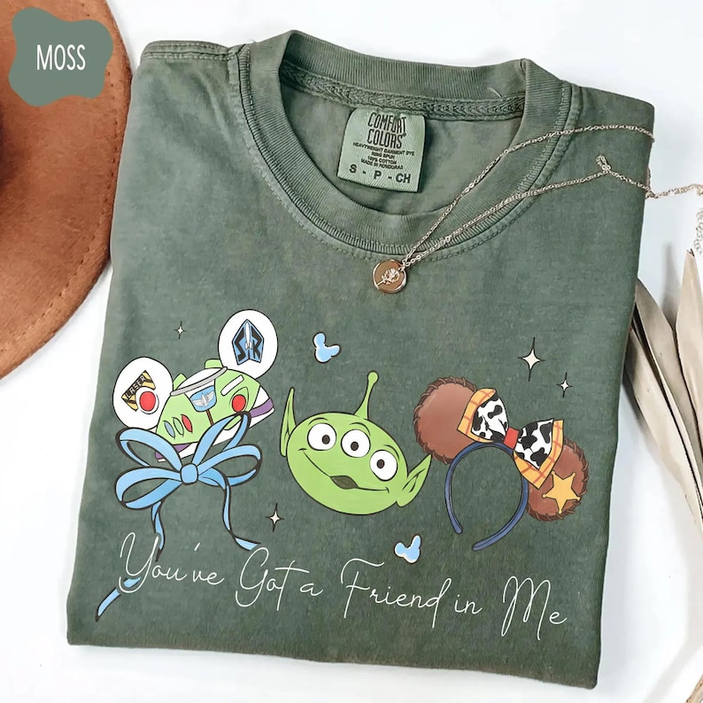 May include: Moss green t-shirt featuring a graphic of Disney characters, including Buzz Lightyear, an alien, and Mickey Mouse ears. The phrase "You've Got a Friend in Me" is written in cursive. A silver necklace is draped across the shirt.