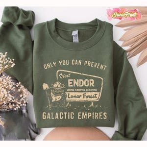 Puede incluir: Una sudadera verde oliva con el texto "ONLY YOU CAN PREVENT Visit ENDOR Hiking-Camping-Feasting Lunar Forest GALACTIC EMPIRES" en un estilo vintage. El diseño presenta un personaje de dibujos animados con una pala y un letrero.