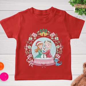 May include: A red t-shirt featuring a snow globe design with two cartoon characters hugging Olaf the snowman. The snow globe is surrounded by a floral wreath with bells and a bow. A small blue dragon is also present.