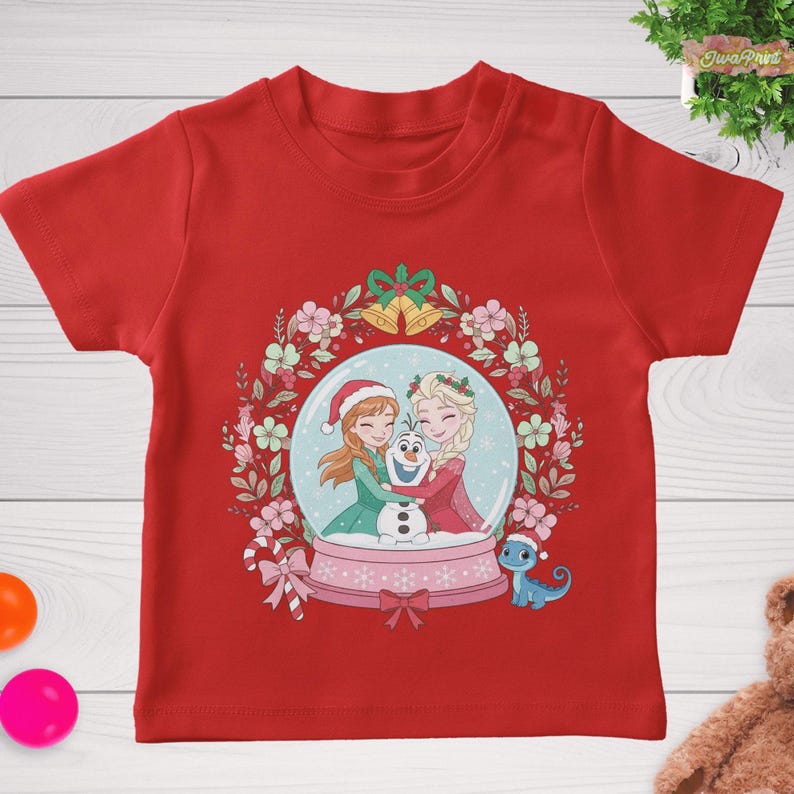 May include: A red t-shirt featuring a snow globe design with two cartoon characters hugging Olaf the snowman. The snow globe is surrounded by a floral wreath with bells and a bow. A small blue dragon is also present.