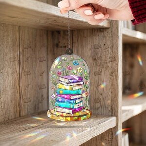 May include: A decorative hanging ornament featuring a stack of colorful books inside a glass dome. The books have purple, yellow, and blue covers. The dome is adorned with floral and leaf designs. The ornament is suspended by a silver chain.