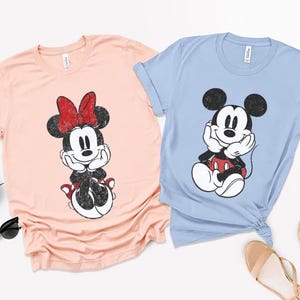 Pode incluir: Duas camisetas, uma p&ecirc;ssego e outra azul claro, cada uma com um gr&aacute;fico desgastado de um rato de desenho animado. A camiseta p&ecirc;ssego tem um la&ccedil;o vermelho e a camiseta azul claro tem um gr&aacute;fico de rato em preto e branco. Ambas as camisetas t&ecirc;m gola redonda.