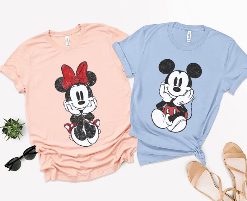 Pode incluir: Duas camisetas, uma p&ecirc;ssego e outra azul claro, cada uma com um gr&aacute;fico desgastado de um rato de desenho animado. A camiseta p&ecirc;ssego tem um la&ccedil;o vermelho e a camiseta azul claro tem um gr&aacute;fico de rato em preto e branco. Ambas as camisetas t&ecirc;m gola redonda.