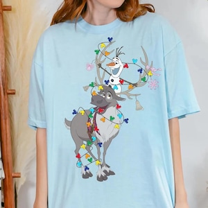 May include: A light blue t-shirt featuring a graphic of a grey reindeer and a white snowman tangled in a string of colorful holiday lights shaped like mouse ears. The reindeer has large antlers, and the snowman has a carrot nose and twig arms.