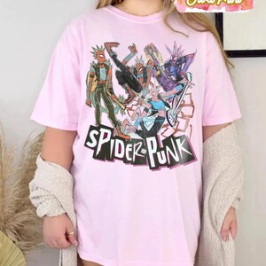 May include: A light pink t-shirt featuring a graphic of several Spider-Man characters in a punk rock style. The words "SPIDER PUNK" are printed in a bold, black font with a white outline. The shirt has short sleeves.