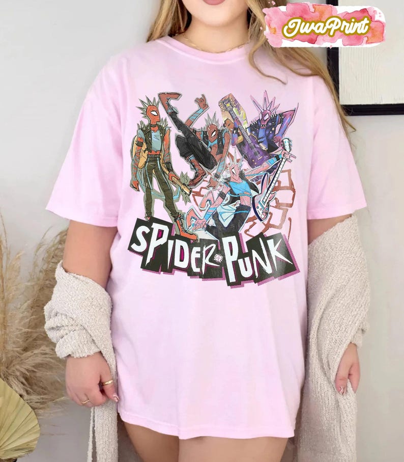 May include: A light pink t-shirt featuring a graphic of several Spider-Man characters in a punk rock style. The words "SPIDER PUNK" are printed in a bold, black font with a white outline. The shirt has short sleeves.