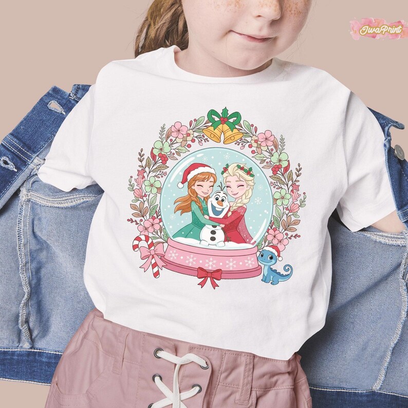 May include: White t-shirt featuring a snow globe design with Anna, Elsa, and Olaf. The snow globe is surrounded by a floral wreath with bells and a candy cane. A small blue dragon wearing a Santa hat is also featured.