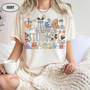 Può includere: T-shirt color avorio con una grafica colorata delle attrazioni e dei personaggi di Hollywood Studios. Il design include il testo "Hollywood Studios", "Toy Story" e "Star Wars". La maglietta è a maniche corte e ha una vestibilità ampia.