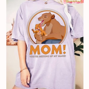 May include: A light purple t-shirt featuring a cartoon lioness and cub inside a circular frame. The text "MOM! YOU'RE MESSING UP MY MANE!" is printed in orange below the image. The shirt has short sleeves.