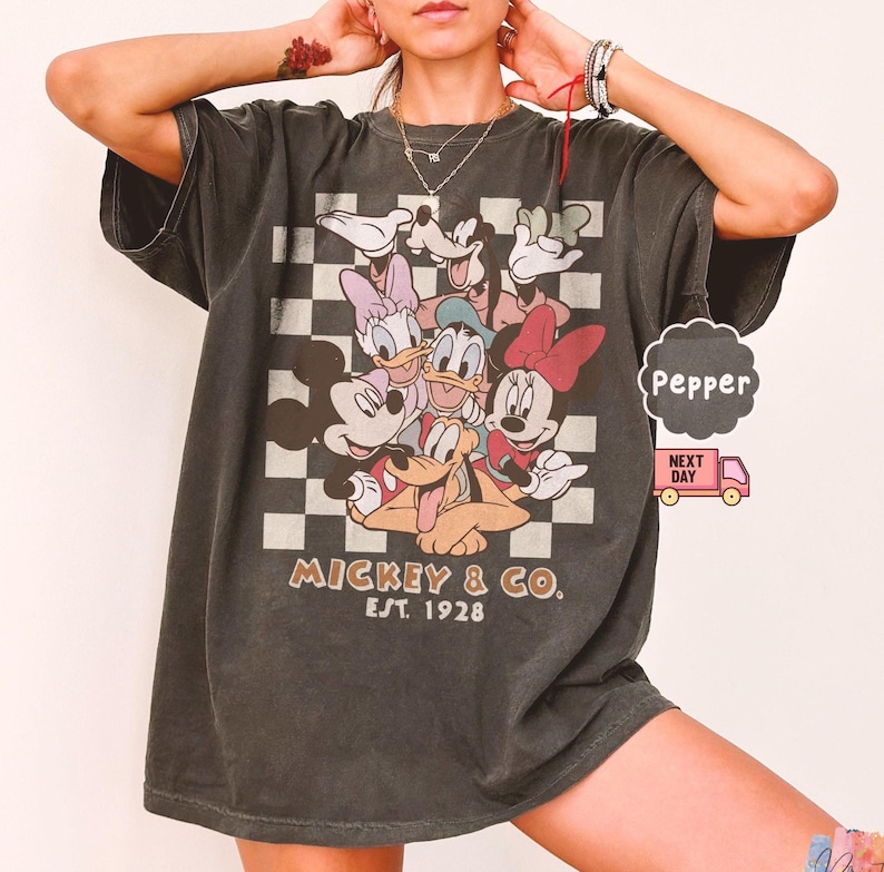May include: Dark gray t-shirt featuring a graphic of Mickey Mouse and friends on a checkered background. The shirt has the text "Mickey & Co. Est. 1928" printed on the front. The shirt is a relaxed fit.