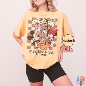 May include: Mustard-colored t-shirt featuring a graphic of Mickey Mouse and friends. The design includes Mickey, Minnie, Donald, Daisy, Goofy, and Pluto, set against a brown and tan checkered background. The text "Mickey & Co. Est. 1928" is printed below the characters.