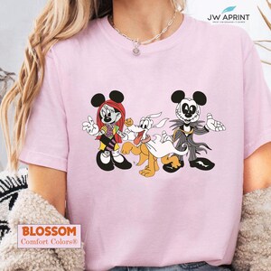 Puede incluir: Camiseta rosa claro con un dibujo animado de Mickey Mouse, Minnie Mouse y Pluto, dise&ntilde;ados como personajes de Pesadilla antes de Navidad. La camiseta tiene el texto "BLOSSOM Comfort Colors&reg;" en la parte inferior.