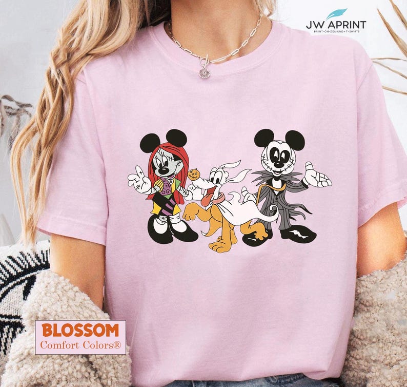 Puede incluir: Camiseta rosa claro con un dibujo animado de Mickey Mouse, Minnie Mouse y Pluto, dise&ntilde;ados como personajes de Pesadilla antes de Navidad. La camiseta tiene el texto "BLOSSOM Comfort Colors&reg;" en la parte inferior.