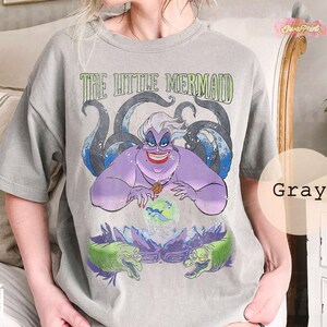 May include: Gray t-shirt featuring a graphic of Ursula from The Little Mermaid. The design includes the text "THE LITTLE MERMAID" above Ursula, with tentacles and two green eels. The shirt is a unisex style.