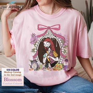 May include: A light pink t-shirt featuring a framed illustration of a character with red hair and a patchwork dress. The frame is decorated with flowers and a pink bow. The shirt also has the text "Blossom" and "Comfort Colors&reg;".