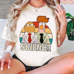 May include: Cream-colored t-shirt featuring a retro-style graphic of two cartoon characters in lab coats, with the word "SCIENCE" in bold letters. The graphic has a vintage, distressed look with a colorful sunset background.