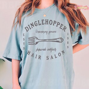 Puede incluir: Camiseta azul claro con un dise&ntilde;o circular que dice "DINGLEHOPPER HAIR SALON" con una ilustraci&oacute;n de tenedor. El texto incluye "Repurposing genuine", "shipwrek artifacts" y "ESTD 1989".